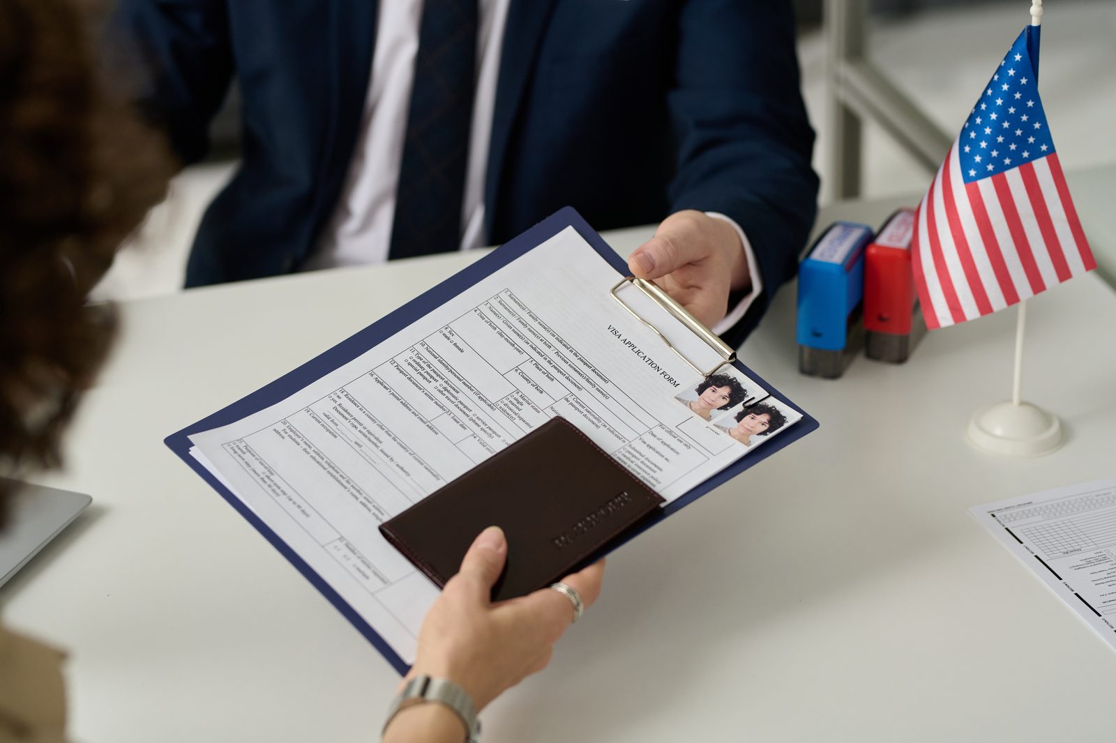 An applicant handing a visa application and passport across an immigration officer's desk, with a small American flag in view